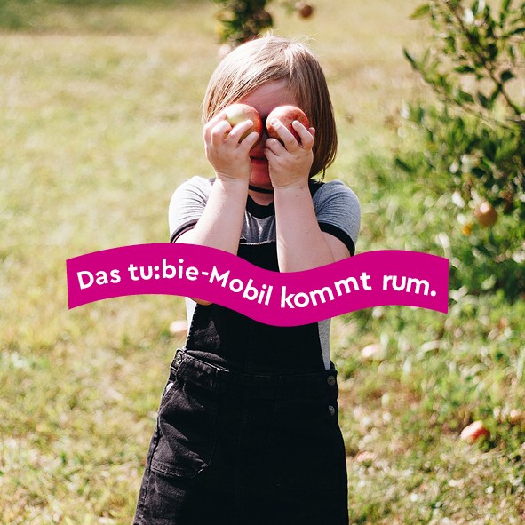 Mädchen hät sich Äpfel vor die Augen und steht auf einer Wiese