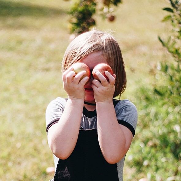 Mädchen hät sich Äpfel vor die Augen und steht auf einer Wiese
