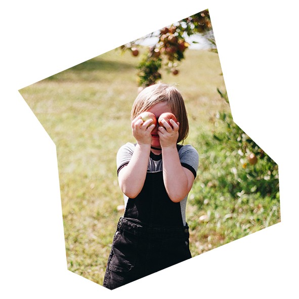 Mädchen hät sich Äpfel vor die Augen und steht auf einer Wiese