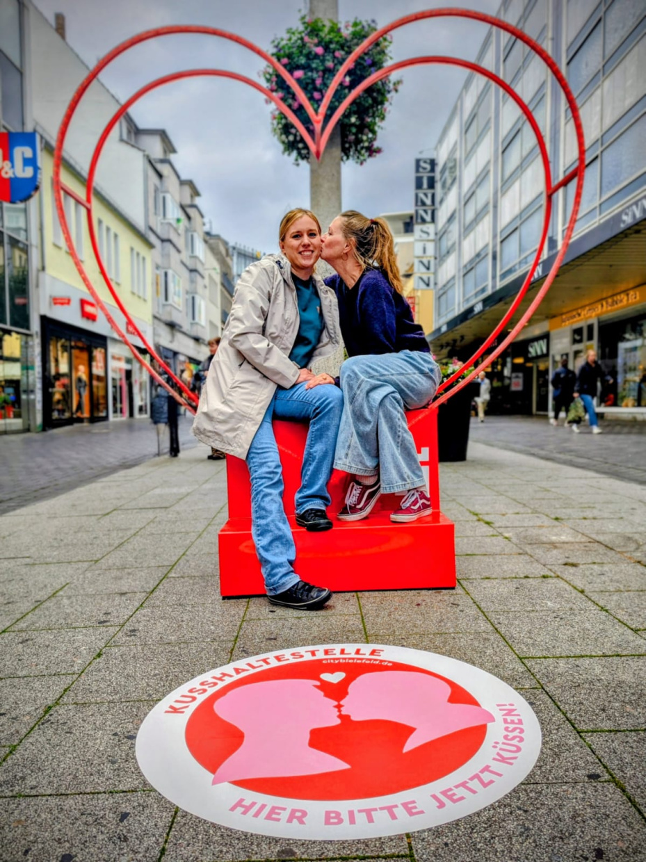 zwei Frauen sitzen vor einem großen Metallherz. Die eine küsst der anderen auf die Wange