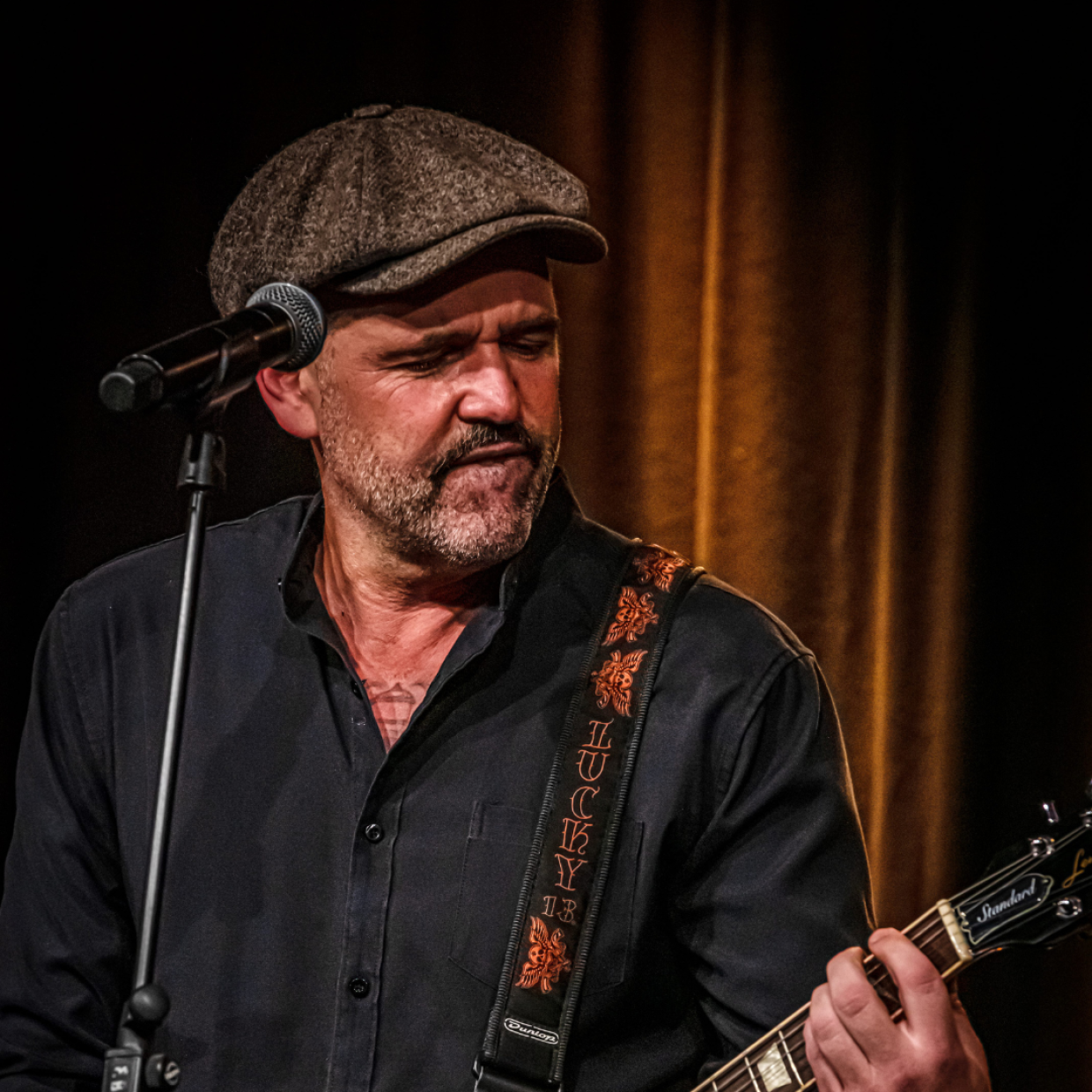Ein Mann der auf der Bühne vor einem Mikro steht und eine Gitarre in der Hand hält und spielt.