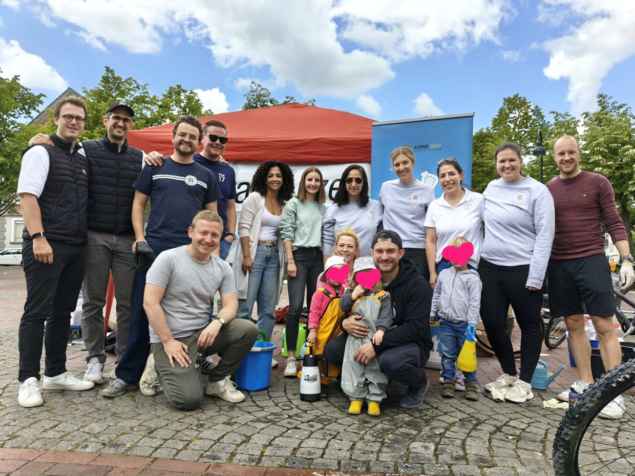 Gruppe von Menschen unterschiedlichen Alters vor einem roten Haus mit blauem Himmel.