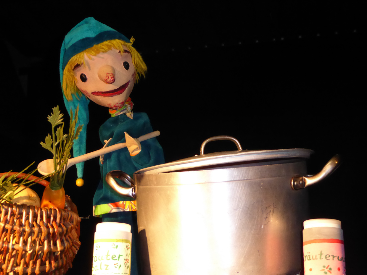 Kasper-Puppe in blauem Outfit neben großem Kochtopf und Gemüse.