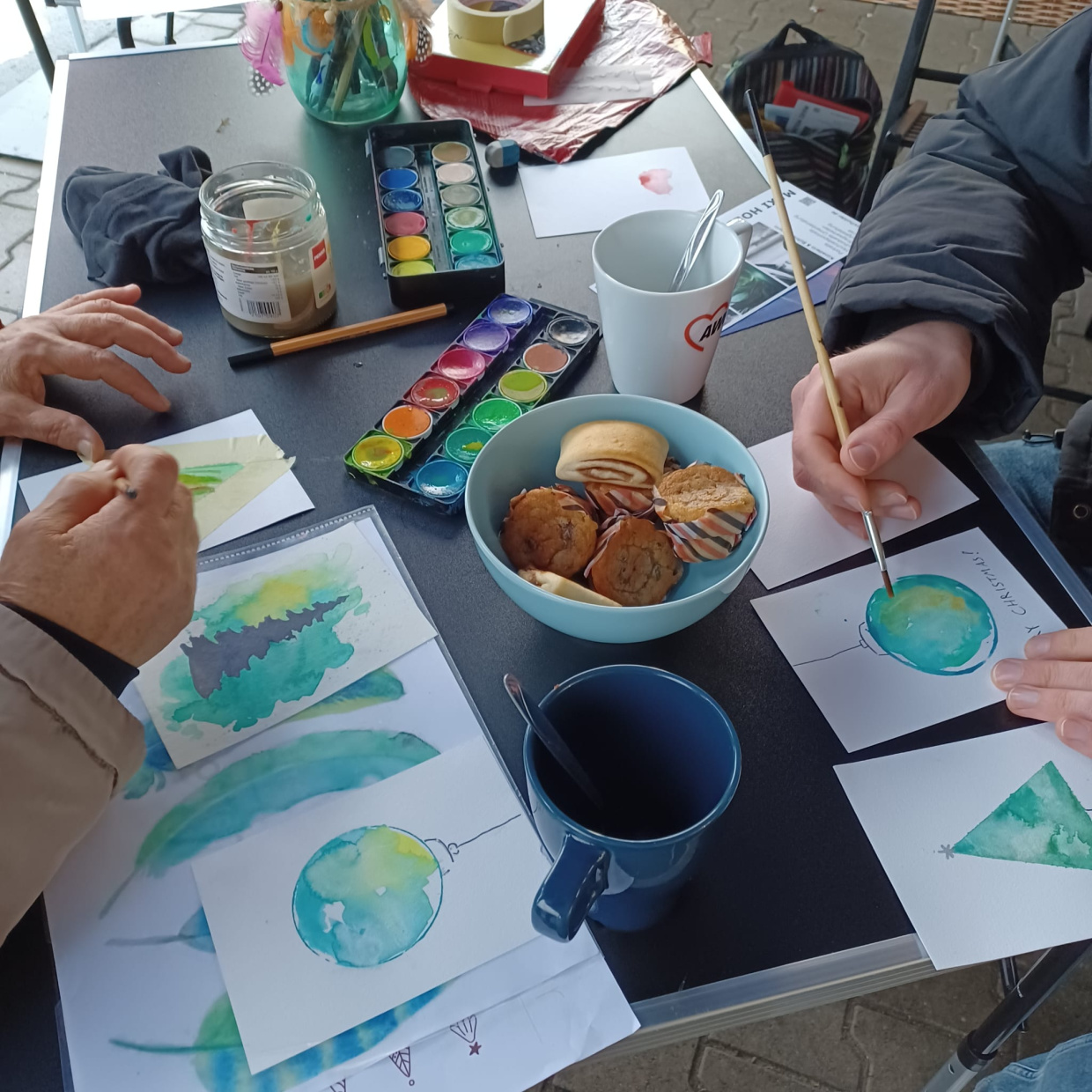 zwei Personen bemalen eine Karte mit Weihnachtsbäumen und Weihnachtskugeln