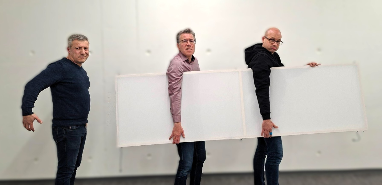 Zwei Menschen tragen eine Kulisse, ein Dritter tut pantomimisch so als ob er mittragen würde.