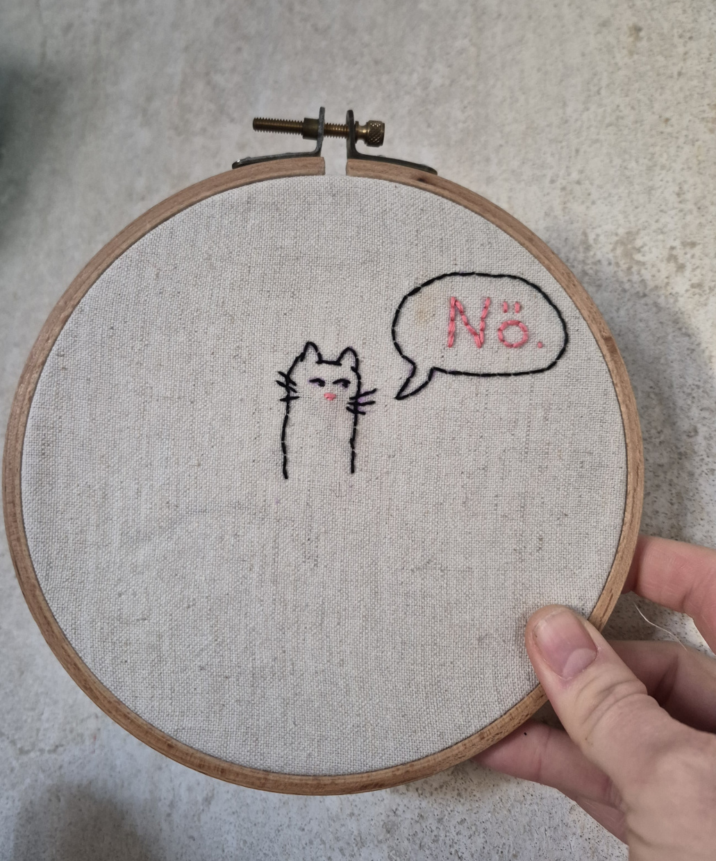 Hand hält Stickrahmen. Auf dem gestickten Bild ist eine Katze mit Sprechblase "Nö." zu sehen.