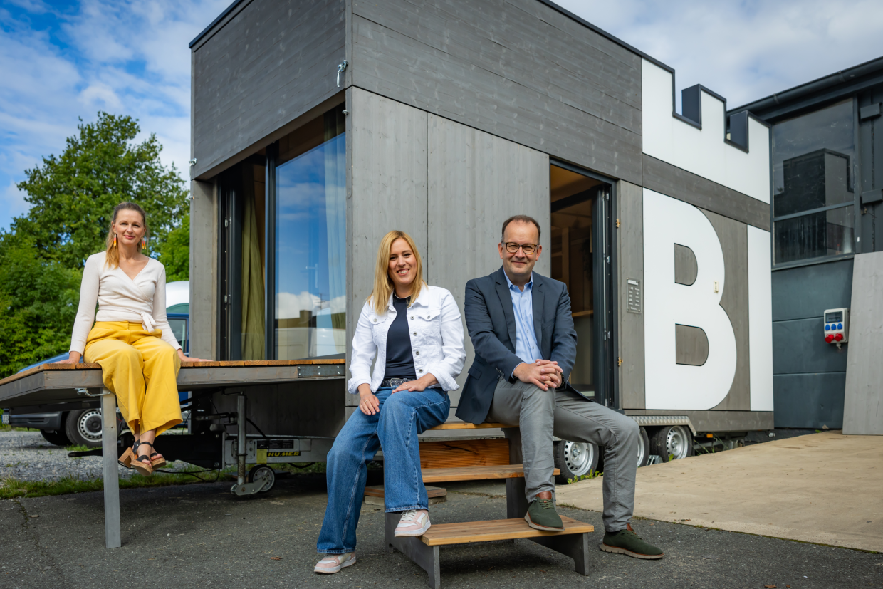 zwei Frauen und ein Mann sitzen vor einem sogenannten Tiny-House