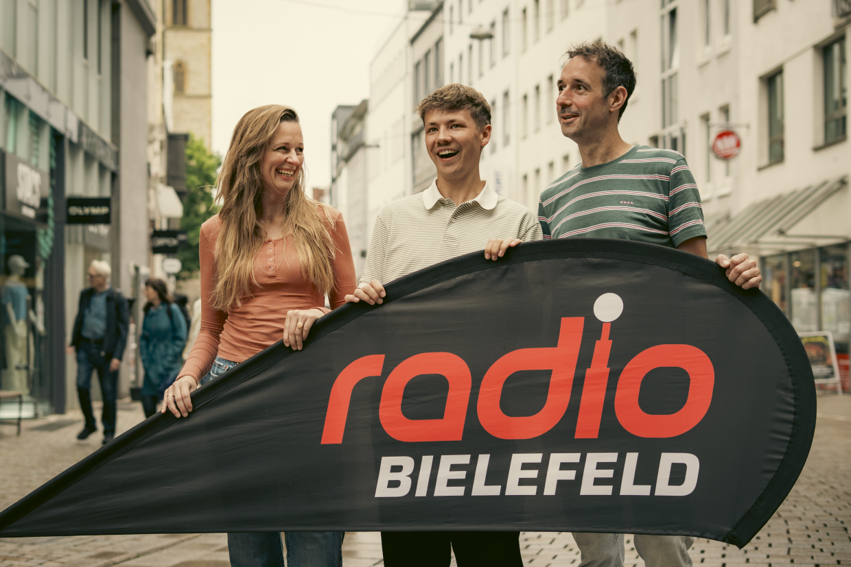 eine Frau und zwei Männer stehen hinter einer Beachflag mit der Aufschrift Radio Bielefeld