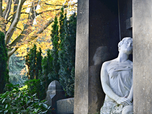 Eine Steinstatue neben einem Gebäude auf dem Johannisfriedhof, im Hintergrund sind verschiedene Pflanzen zu sehen.