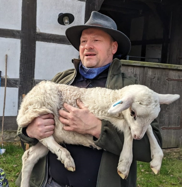 Schäfer Thomas Rüping steht vor dem alten Schafstall auf Hof Ramsbrock und trägt ein weißes Lamm auf dem Arm