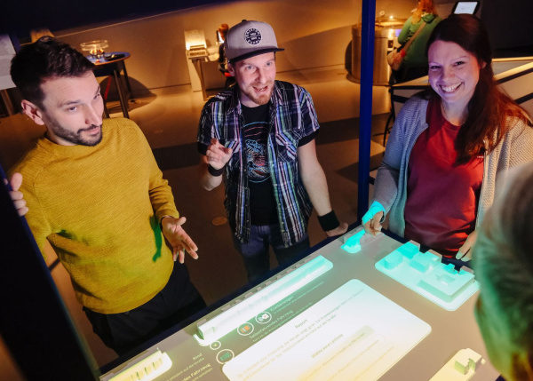 Drei junge Erwachsene experimentieren gemeinsam an einem interaktiven Multitouch-Tisch in einer Ausstellung