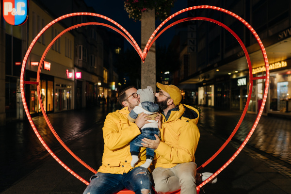 zwei Männer küssen ein Baby auf die Wange vor einem Herzrahmen