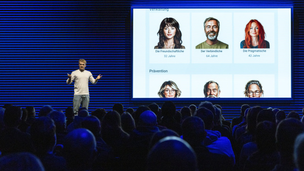 ein Redner steht auf der Bühne, im Vordergrund sitzendes Publikum, neben dem Redner eine Präsentation, die verschiedene Menschenköpfe zeigt