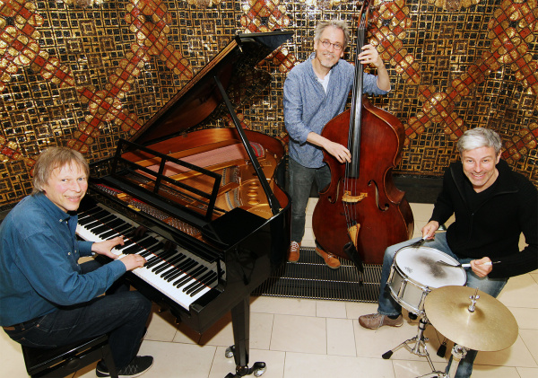 Drei Musiker spielen zusammen: Klavier, Cello und Schlagzeug in einem Raum mit gemusterter Wand.