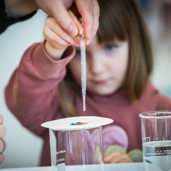 Auf dem Bild ist Hintergrund ein Kind unscharf zu erkennen. Im Vordergrund hält es eine Pipette in der Hand hält und auf ein Tuch etwas tröpfeln lässt.