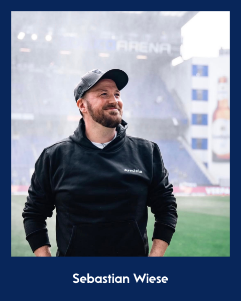 Sebastian Wiese trägt schwarze Jacke und Kappe, Stadion-Hintergrund.