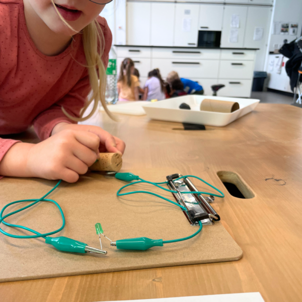 Ein Bild von einem Kind welches auf einen Tisch lehnt, vor ihr ein Klemmbrett mit Kabeln die einen Stromkreislauf schließen.