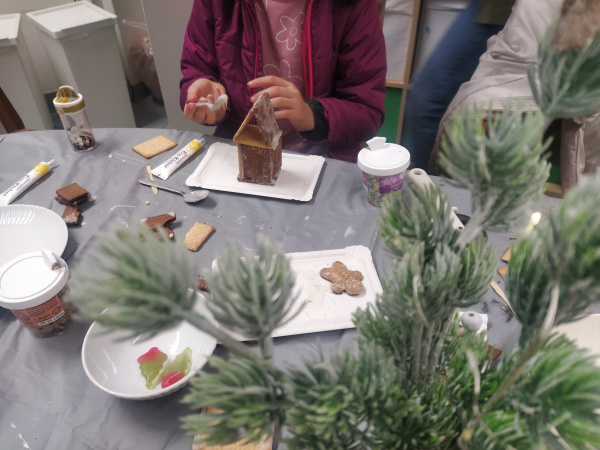 ein Haus aus Lebkuchen mit Zuckerguss wird gebastelt, davor sind Tannenzweige zu sehen