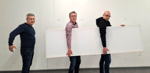 Zwei Menschen tragen eine Kulisse, ein Dritter tut pantomimisch so als ob er mittragen würde.