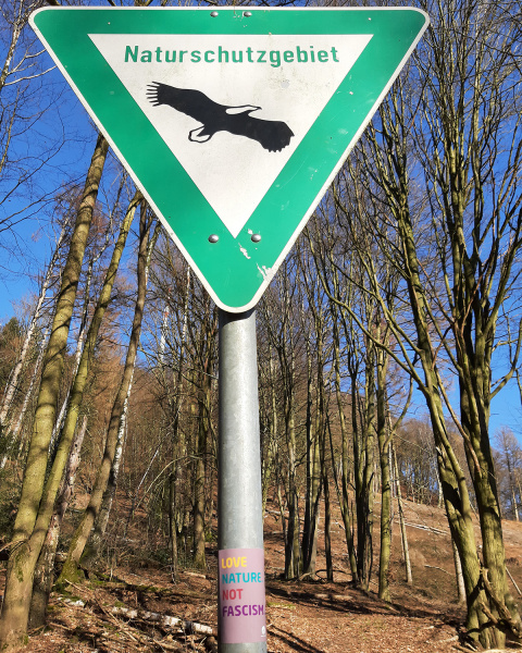 Zu sehen ist ein Schild für ein Naturschutzgebie. An dem Pfeiler steht "Love Nature not Fascism"