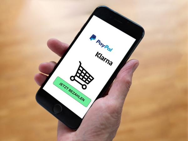 Smartphone-Display zeigt PayPal- und Klarna-Logo mit Einkaufswagen-Symbol und grünem Bezahl-Button