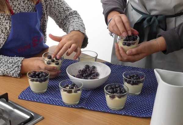 Zwei Kinder mit Schürzen garnieren kleine Gläser mit Vanillecreme und frischen Heidelbeeren.