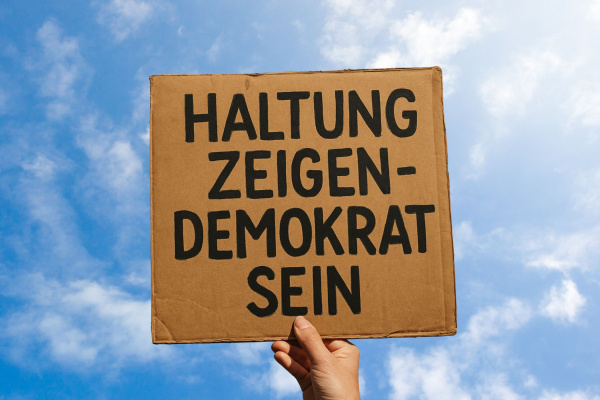 Eine Hand hält ein Plakat mit der Aufschrift "Haltung zeigen - Demokrat sein" in den blauen Himmel. 