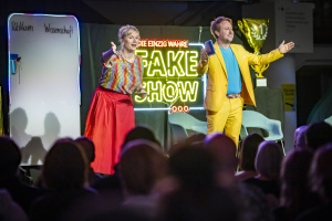 Zwei Moderator:innen stehen auf einer Bühne vor Publikum. Im Hintergrund leuchtet ein buntes „FAKE SHOW“-Schild. Links eine Tafel mit den Rubriken „Publikum“ und „Wissenschaft“. Die Moderatorin gestikuliert lebhaft, der Moderator im knallgelben Anzug hält eine Karte in der Hand.