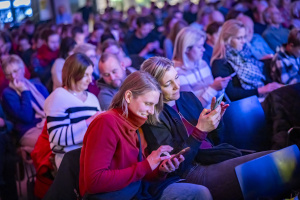 Zuschauer:innen in einem voll besetzten Veranstaltungsraum, viele mit Blick auf ihre Smartphones. Im Vordergrund lachen zwei Frauen, während sie gemeinsam auf ihre Handys schauen. Die Szene ist bunt beleuchtet und zeigt ein konzentriertes, interaktives Publikum. ChatGPT fragen