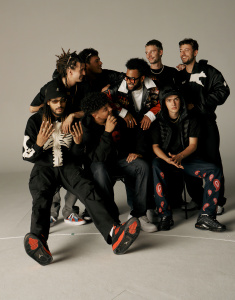 Gruppe von acht Personen in schwarzer Kleidung posiert zusammen vor neutralem Hintergrund.